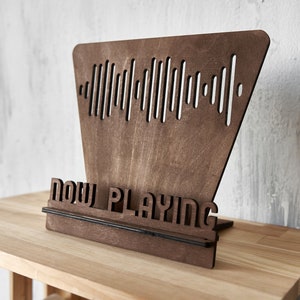 May include: A wooden stand with a cutout of a sound wave and the words "NOW PLAYING" on the front. The stand is made of dark brown wood and has a black metal bar across the front.