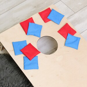 Puede incluir: Un tablero de cornhole de madera de color claro con un agujero circular en el centro, acompa&ntilde;ado de ocho bolsas de frijoles cuadradas en colores rojo y azul alternos dispersas por la superficie del tablero para un juego de jard&iacute;n.