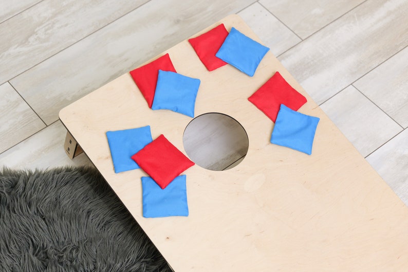 Puede incluir: Un tablero de cornhole de madera de color claro con un agujero circular en el centro, acompa&ntilde;ado de ocho bolsas de frijoles cuadradas en colores rojo y azul alternos dispersas por la superficie del tablero para un juego de jard&iacute;n.