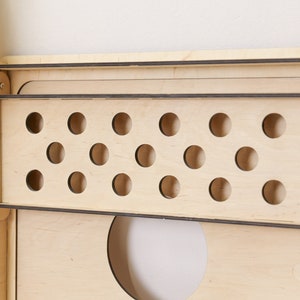 Puede incluir: Un organizador de pared de madera clara que cuenta con un estante rectangular con un panel frontal que contiene dos filas horizontales de recortes circulares. La estructura est&aacute; construida con l&aacute;minas de madera contrachapada con bordes oscuros.