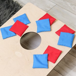 Puede incluir: Un tablero de cornhole de madera de color claro con un agujero circular en el centro, acompa&ntilde;ado de cuatro bolsas de frijoles de tela roja y cuatro bolsas de frijoles de tela azul esparcidas por la superficie plana del tablero para el juego.