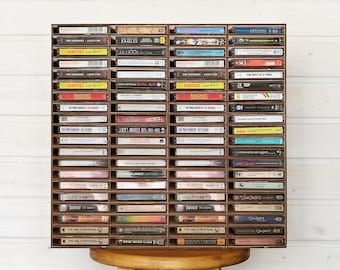 Könnte beinhalten: Ein braunes Holzregal mit vier vertikalen Spalten, die jeweils mehrere horizontale Fächer enthalten, welche mit verschiedenen Musikkassetten gefüllt sind. Das Regal ist so organisiert, dass es zahlreiche Kassetten aufnimmt und deren Rücken mit Titeln zeigt.
