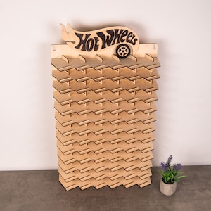 May include: A light-coloured wooden wall-mounted display rack featuring a Hot Wheels logo at the top with a cutout wheel design. The rack has multiple horizontal shelves with notched edges, designed to hold small toy cars in a vertical arrangement.
