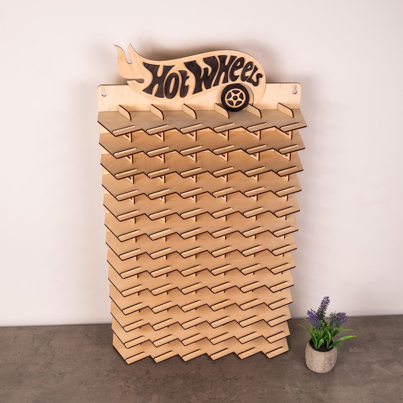 May include: A light-coloured wooden wall-mounted display rack featuring a Hot Wheels logo at the top with a cutout wheel design. The rack has multiple horizontal shelves with notched edges, designed to hold small toy cars in a vertical arrangement.