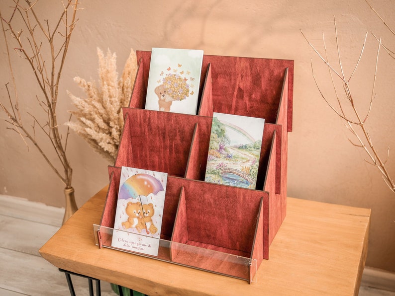 K&ouml;nnte beinhalten: Ein roter Kartenst&auml;nder aus Holz mit f&uuml;nf F&auml;chern zum Aufbewahren von Gru&szlig;karten. Der St&auml;nder hat eine transparente Acrylglasscheibe vorne und ist mit Karten gef&uuml;llt, die verschiedene Illustrationen zeigen.