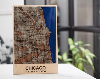 Pode incluir: Um mapa de madeira de Chicago com um enchimento de epóxi azul para o Lago Michigan. O mapa é cortado a laser e apresenta as ruas e os bairros da cidade. O texto "CHICAGO" e as coordenadas "41°52'55"N 87°37'40"W" estão impressas abaixo do mapa.