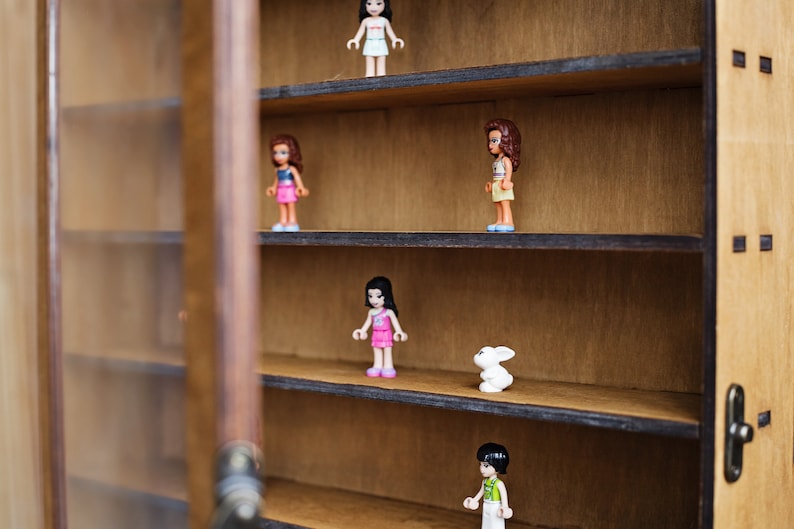 May include: A wooden display case with five shelves, each holding a small plastic figurine. The figurines are of people, including a girl with brown hair and a pink dress, a girl with brown hair and a blue dress, a girl with brown hair and a yellow dress, a girl with brown hair and a white dress, and a boy with brown hair and a green shirt. There is also a small white rabbit figurine on one of the shelves.