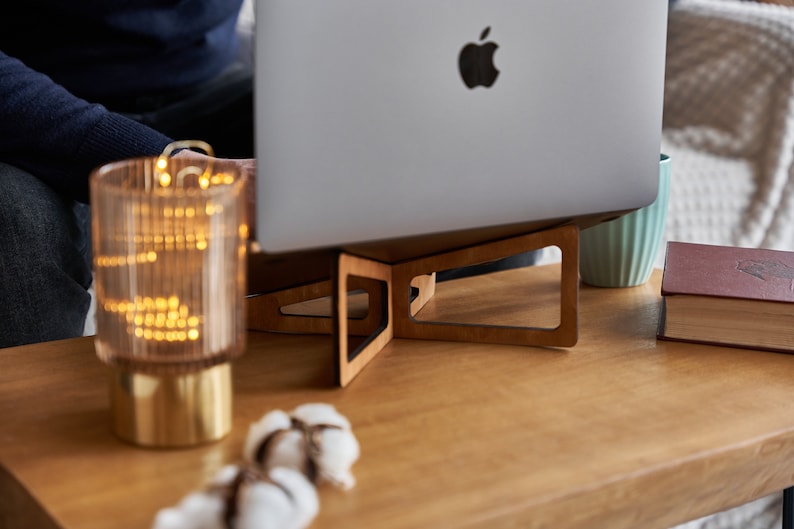 K&ouml;nnte beinhalten: Ein Laptopst&auml;nder aus Holz mit hellbrauner Oberfl&auml;che und geometrischem Design. Der St&auml;nder h&auml;lt einen silbernen Laptop mit einem Apple-Logo. Der Laptop steht auf einem Holztisch mit einer Glaslampe und Watteb&auml;uschen.