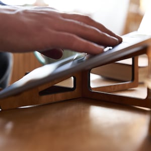 K&ouml;nnte beinhalten: Ein Laptop-St&auml;nder aus Holz mit dunkelbrauner Oberfl&auml;che. Der St&auml;nder ist angewinkelt, um einen Laptop oder ein Tablet zu erh&ouml;hen. Der St&auml;nder steht auf einem Holztisch.