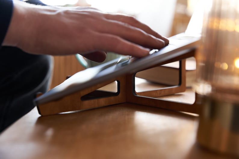 K&ouml;nnte beinhalten: Ein Laptop-St&auml;nder aus Holz mit dunkelbrauner Oberfl&auml;che. Der St&auml;nder ist angewinkelt, um einen Laptop oder ein Tablet zu erh&ouml;hen. Der St&auml;nder steht auf einem Holztisch.
