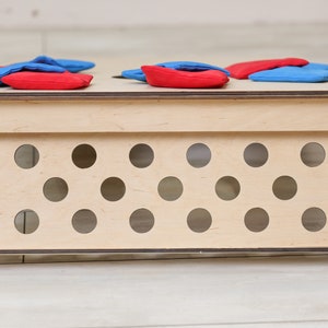 Puede incluir: Un tablero de juego de madera rectangular con un acabado de madera natural clara y bordes oscuros. El panel frontal est&aacute; perforado con doce recortes circulares dispuestos en dos filas. Varias bolsas de tela rojas y azules descansan sobre la superficie.