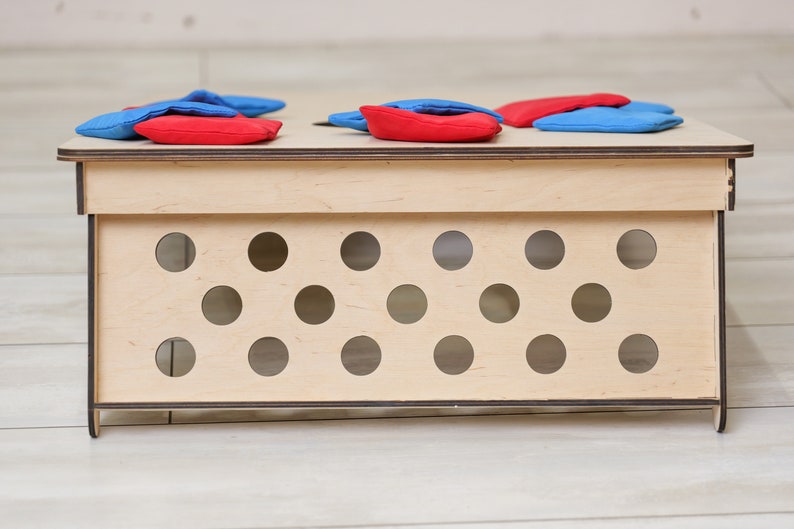 Puede incluir: Un tablero de juego de madera rectangular con un acabado de madera natural clara y bordes oscuros. El panel frontal est&aacute; perforado con doce recortes circulares dispuestos en dos filas. Varias bolsas de tela rojas y azules descansan sobre la superficie.