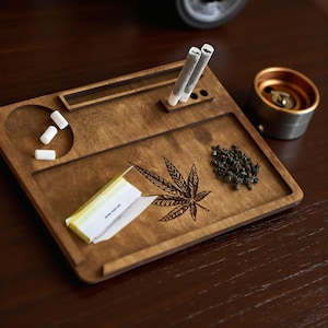 Può includere: Vassoio in legno per rollare con un design a foglia di cannabis intagliata. Il vassoio ha una sezione rotonda con filtri bianchi, una sezione per cartine e una sezione con due sigarette prerollate. Sono presenti anche un piccolo mucchio di erbe verdi e un posacenere in metallo.