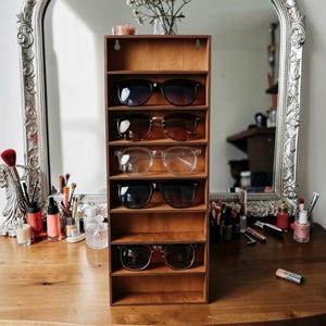 Peut inclure: Un présentoir vertical en bois doté de sept étagères ouvertes, conçu pour être fixé au mur. Le support contient cinq paires de lunettes, dont des lunettes de soleil teintées foncées et une paire à monture transparente, disposées sur des niveaux.