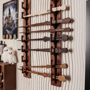 Op de afbeelding: Een verticaal houten wandrek met twee parallelle donkerbruine planken met inkepingen die zeven verschillende houten toverstokken vasthouden. Elke toverstok is gegraveerd met tekst, waaronder Expelliarmus en Expecto Patronum, in diverse houttinten.
