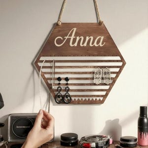 May include: A hexagonal dark wood jewellery organiser hanging by a rope, featuring the name Anna in white script at the top. The board has horizontal slats with small holes for earrings and a scalloped bottom edge, holding black drop earrings.