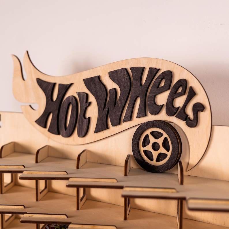 May include: A light-coloured wooden display rack featuring the Hot Wheels logo in dark wood lettering. The sign includes a stylised dark wood wheel graphic integrated into the design. The rack has multiple tiered shelves with dividers for organising small toy cars.