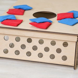 Puede incluir: Un tablero de cornhole de madera clara con un orificio circular en la superficie superior y un panel lateral con m&uacute;ltiples recortes circulares. Varios sacos de tela rojos y azules est&aacute;n esparcidos por la parte superior del tablero.