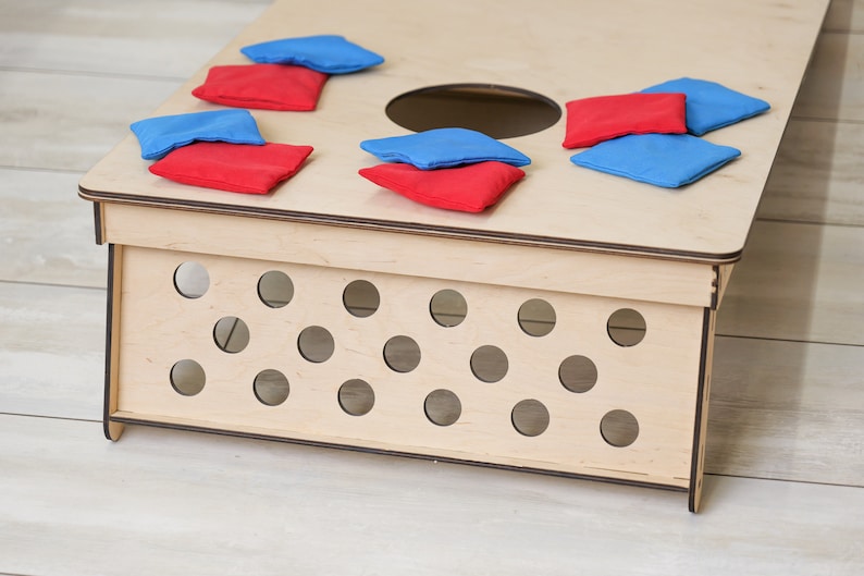 Puede incluir: Un tablero de cornhole de madera clara con un orificio circular en la superficie superior y un panel lateral con m&uacute;ltiples recortes circulares. Varios sacos de tela rojos y azules est&aacute;n esparcidos por la parte superior del tablero.
