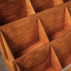 Pu&ograve; includere: Un organizer in legno con pi&ugrave; scomparti. Gli scomparti sono divisi da divisori verticali e angolati, creando una struttura a griglia. Il legno ha una calda tinta marrone-rossastra e la costruzione appare robusta.