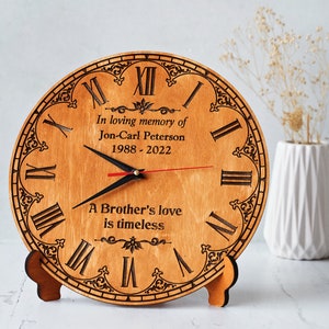 May include: A wooden memorial clock with Roman numerals and decorative accents. The clock face includes the text "In loving memory of Jon-Carl Peterson 1988-2022" and "A Brother's love is timeless". The clock has black hands and a red second hand.