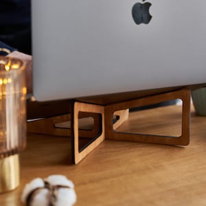 K&ouml;nnte beinhalten: Ein Laptop-St&auml;nder aus Holz mit einer nat&uuml;rlichen Holzoptik und schwarzen Akzenten. Der St&auml;nder ist so konzipiert, dass er einen Laptop f&uuml;r ergonomischen Komfort und eine bessere K&ouml;rperhaltung anhebt.