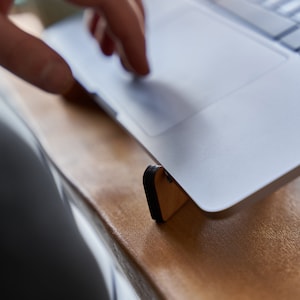 K&ouml;nnte beinhalten: Ein silberner Laptop-Computer steht auf einem Holzst&auml;nder mit schwarzen Gummif&uuml;&szlig;en. Der Laptop liegt auf einer hellbraunen Oberfl&auml;che.