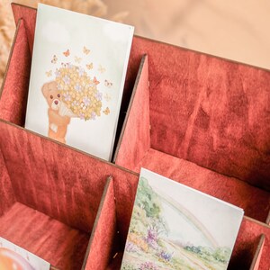 K&ouml;nnte beinhalten: Ein Kartenhalter aus Holz mit sechs F&auml;chern, in einem tiefen Rot lackiert. Der Halter ist mit Karten gef&uuml;llt, die verschiedene Illustrationen zeigen, darunter einen B&auml;ren, der Blumen h&auml;lt, und einen Regenbogen.