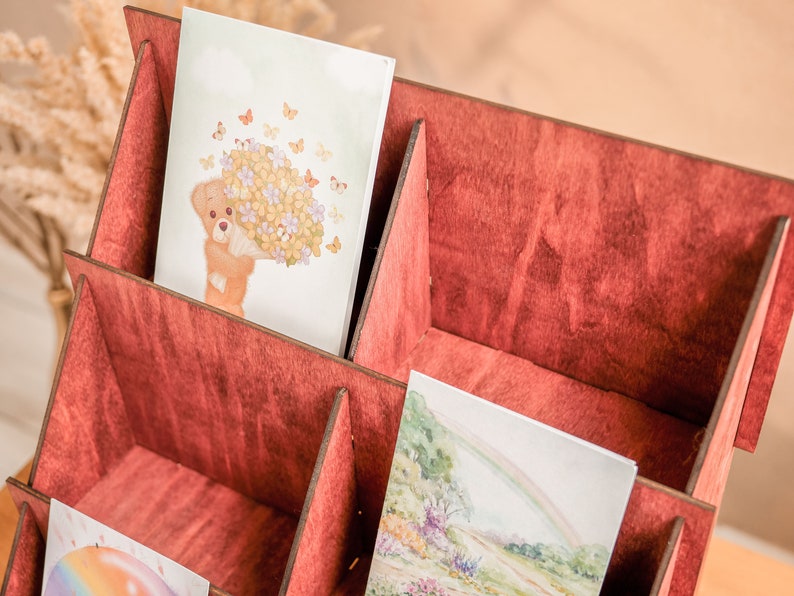 K&ouml;nnte beinhalten: Ein Kartenhalter aus Holz mit sechs F&auml;chern, in einem tiefen Rot lackiert. Der Halter ist mit Karten gef&uuml;llt, die verschiedene Illustrationen zeigen, darunter einen B&auml;ren, der Blumen h&auml;lt, und einen Regenbogen.
