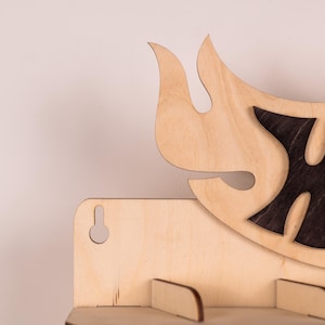 May include: A light-coloured wooden wall shelf featuring a decorative flame-shaped cutout design at the top. The shelf includes a keyhole slot on the left side for mounting and a flat horizontal surface supported by small wooden brackets.