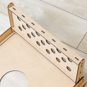 Puede incluir: Un tablero de juego de madera clara con un panel trasero vertical que presenta dos filas de recortes circulares. La estructura est&aacute; ensamblada con juntas visibles y bordes oscuros, mostrando una base plana con una gran abertura circular.