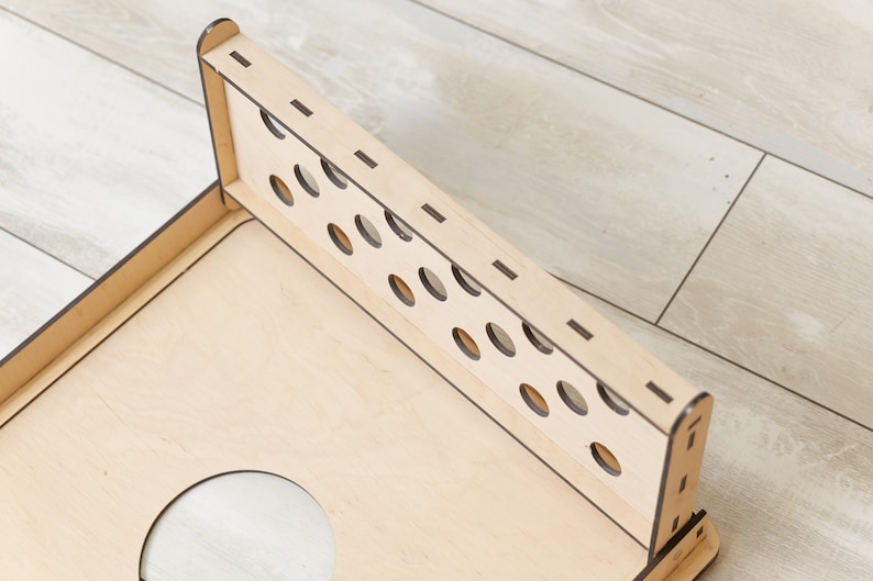 Puede incluir: Un tablero de juego de madera clara con un panel trasero vertical que presenta dos filas de recortes circulares. La estructura est&aacute; ensamblada con juntas visibles y bordes oscuros, mostrando una base plana con una gran abertura circular.
