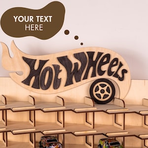 May include: A light-coloured wooden wall-mounted display rack featuring three horizontal shelves for toy cars. The top section displays the dark-stained text Hot Wheels with a wheel icon. A brown speech bubble above contains the text YOUR TEXT HERE.