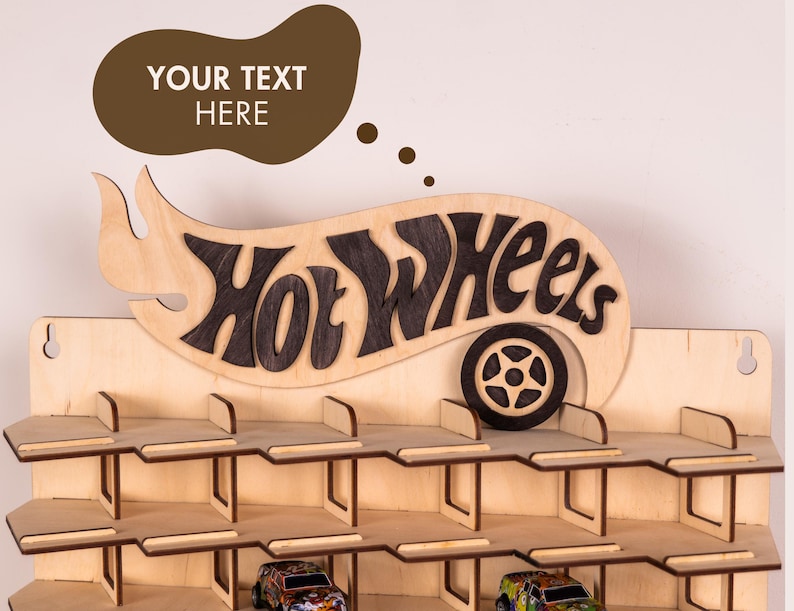 May include: A light-coloured wooden wall-mounted display rack featuring three horizontal shelves for toy cars. The top section displays the dark-stained text Hot Wheels with a wheel icon. A brown speech bubble above contains the text YOUR TEXT HERE.
