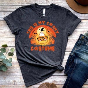 May include: A dark gray t-shirt with a graphic of a candy corn wearing glasses and a witch flying in the background. The text on the shirt reads "This is my corny costume".