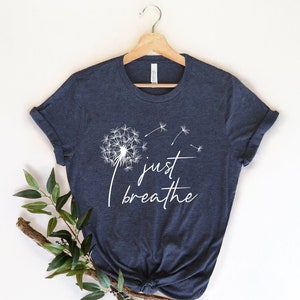 Pu&ograve; includere: Una maglietta blu navy con una grafica di dente di leone bianca e il testo "just breathe".