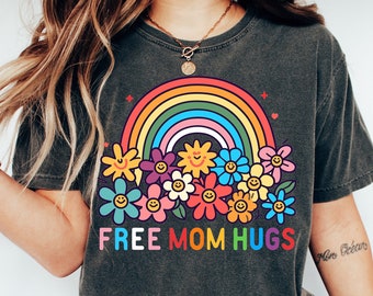 May include: Dark grey t-shirt with a colourful rainbow and flower graphic. The shirt features the text "FREE MOM HUGS" in a playful font. The design includes a rainbow, smiling flowers, and a heart.