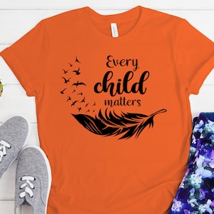 May include: Orange t-shirt with a black feather design and the text "Every child matters".