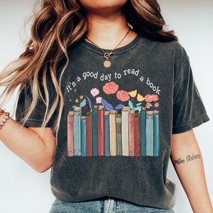 May include: A dark gray t-shirt with a graphic of a stack of colorful books with flowers and the text "It's a good day to read a book".