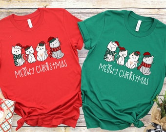 May include: Two Christmas themed t-shirts with a red and green color scheme. The shirts feature a line of five white cats wearing Santa hats and scarves with the text "MEOWY CHRISTMAS".