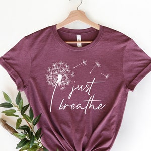 Pu&ograve; includere: Una maglietta color borgogna con un motivo di dente di leone bianco e il testo "just breathe".