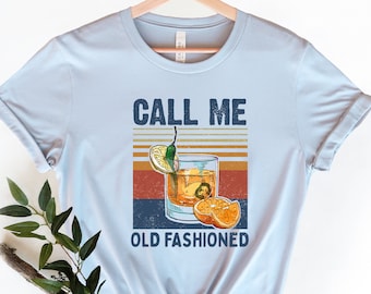 May include: Light blue t-shirt with the text "CALL ME OLD FASHIONED" above a graphic of an Old Fashioned cocktail. The graphic includes a cocktail glass with ice, a lemon slice, and an orange slice.