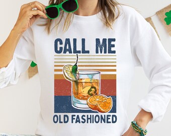 May include: White sweatshirt with a vintage-style graphic of a cocktail glass with orange slices and a green chili pepper. The text "Call Me Old Fashioned" is printed below the image.