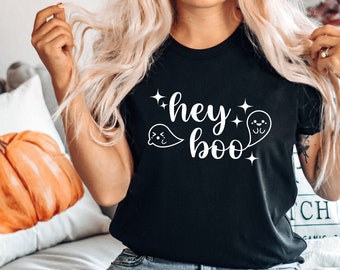 Puede incluir: Una camiseta negra con el texto "hey boo" en letras blancas y dos ilustraciones de fantasmas blancos. La camiseta la lleva puesta una persona con el pelo rubio.