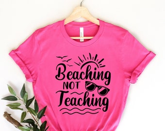 May include: A pink t-shirt with a black graphic that says "Beaching Not Teaching" with a sun, birds, and sunglasses.