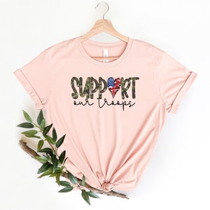 May include: A pink t-shirt with the text "SUPPORT our troops" in black and green camouflage print. A red, white, and blue heart with a camouflage pattern is in the middle of the word "SUPPORT".