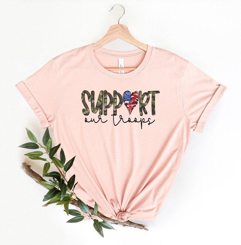 May include: A pink t-shirt with the text "SUPPORT our troops" in black and green camouflage print. A red, white, and blue heart with a camouflage pattern is in the middle of the word "SUPPORT".