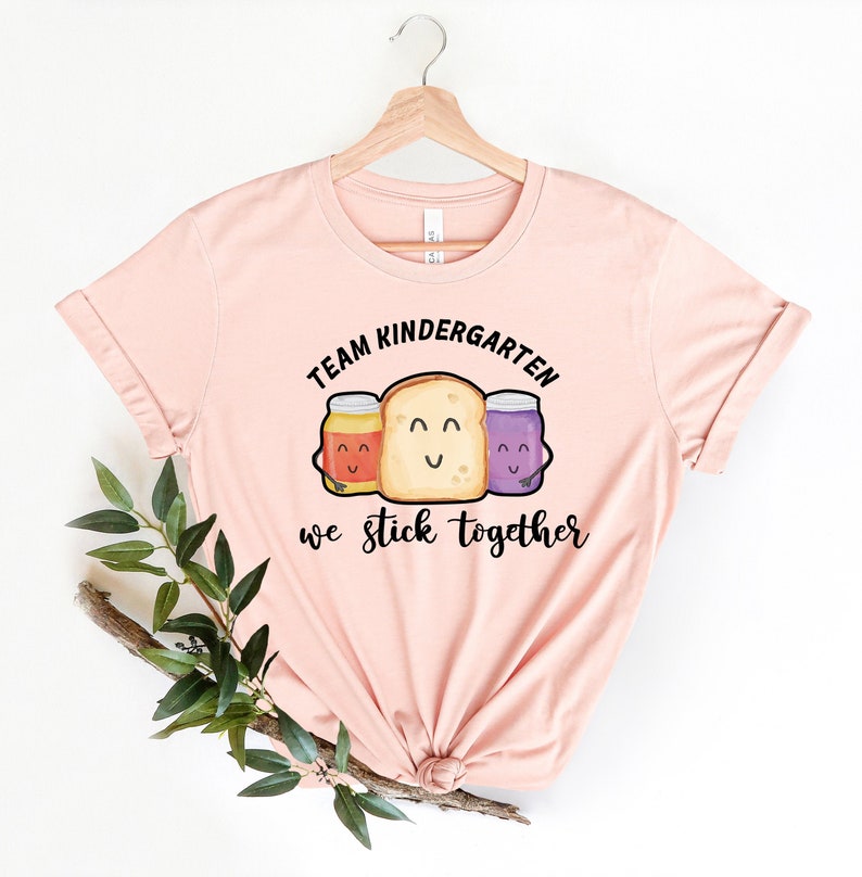 May include: A pink t-shirt with the text "Team Kindergarten we stick together" in black. The text is surrounded by a cartoon image of a slice of bread with two jars of jam on either side.