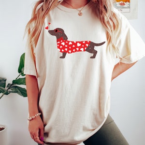 May include: A white t-shirt with a brown dachshund wearing a red sweater with white hearts printed on it. The dachshund is wearing red heart-shaped sunglasses and has two red hearts above its head.