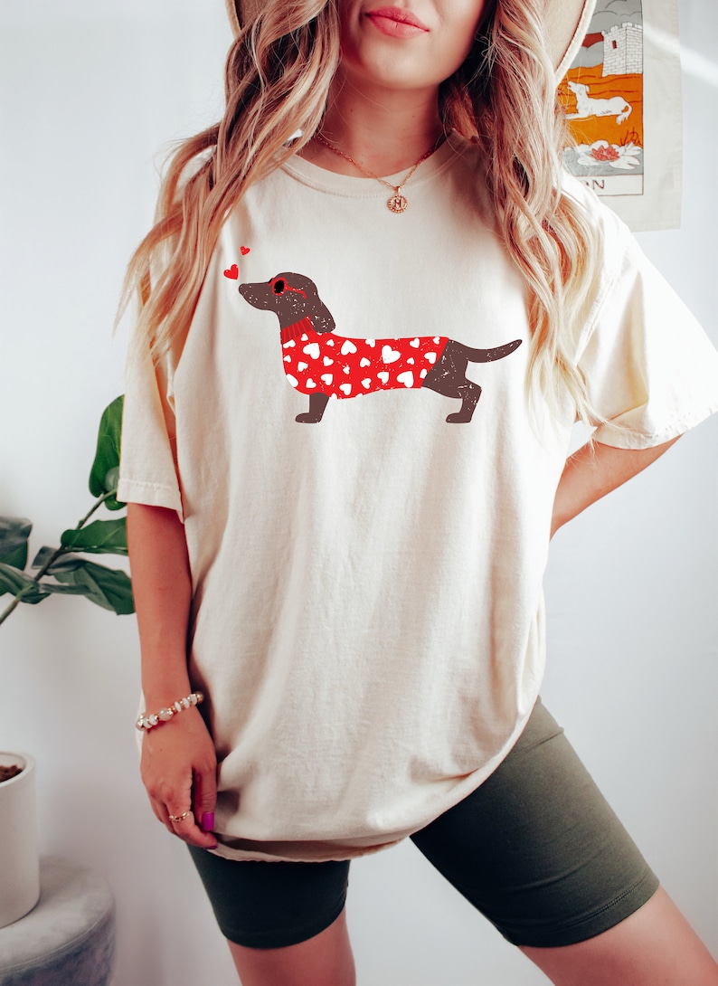 May include: A white t-shirt with a brown dachshund wearing a red sweater with white hearts printed on it. The dachshund is wearing red heart-shaped sunglasses and has two red hearts above its head.